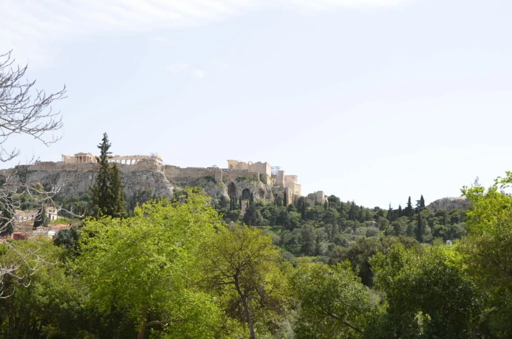 Ancient citadel ruins perched on a hilltop surrounded by green vegetation and cypress trees under a light blue sky