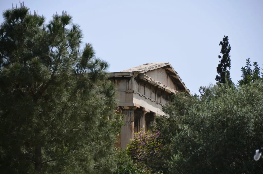 Ancient Greek temple structure with classical columns partially obscured by surrounding trees and vegetation