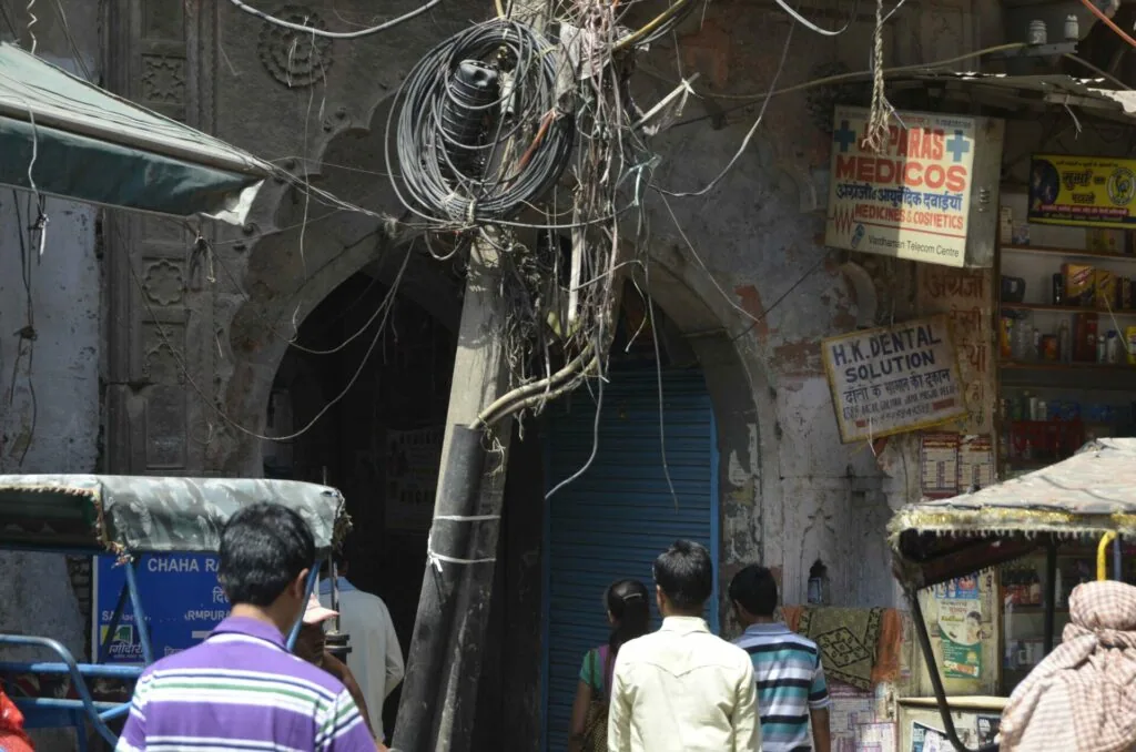 Crowded urban alleyway with tangled overhead electrical wires, weathered building facade, business signs in local script, and pedestrians walking.