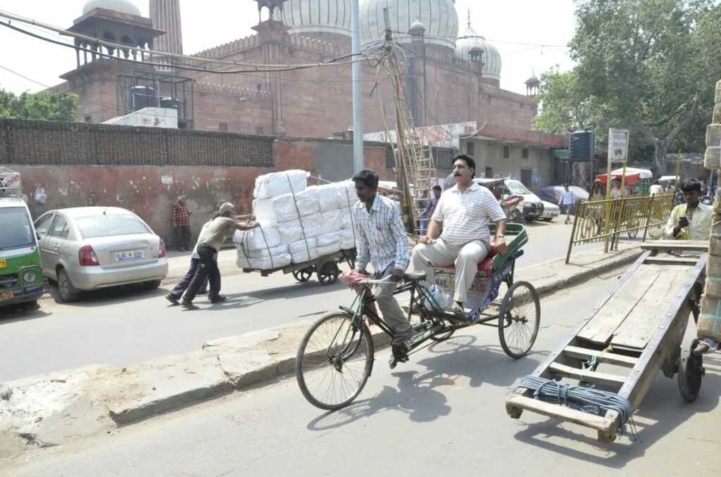 Man riding cargo bicycle loaded with large white wrapped bundle on crowded street with historic brick building and domed structure in background