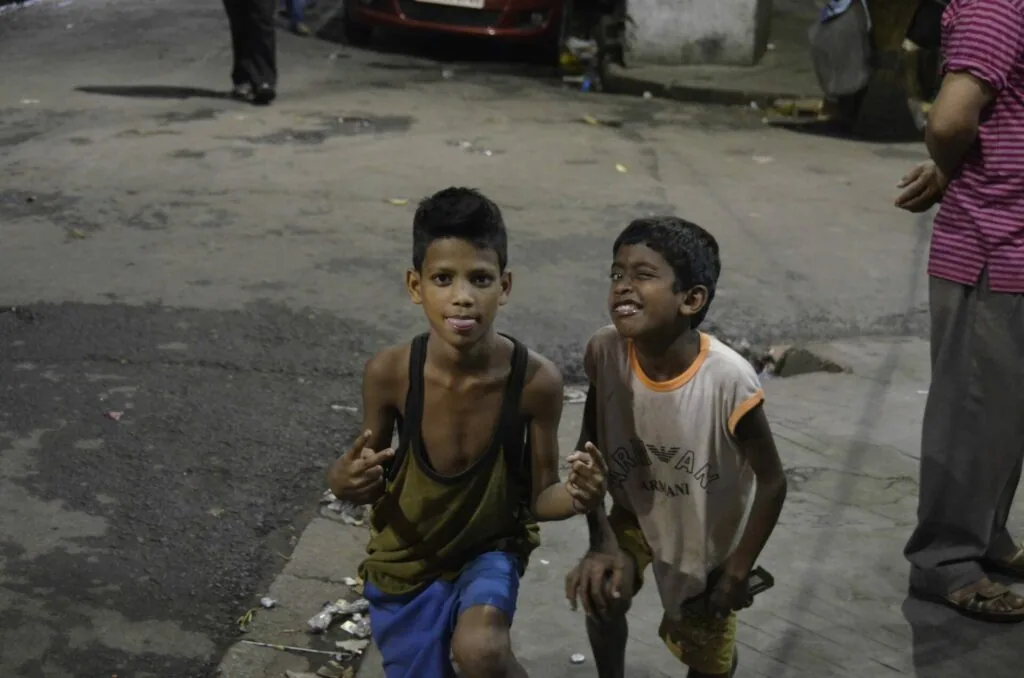 Two young boys smiling while running on a city street with parked cars and people in the background
