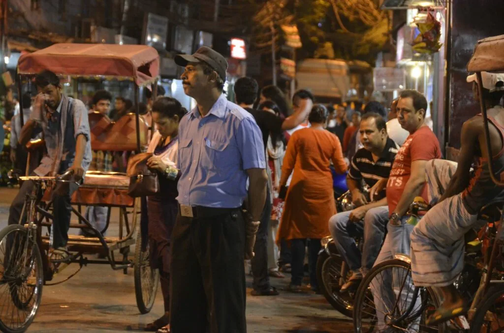 Street scene at night with vendor in blue shirt operating cycle rickshaw and crowds of people walking through busy urban market area