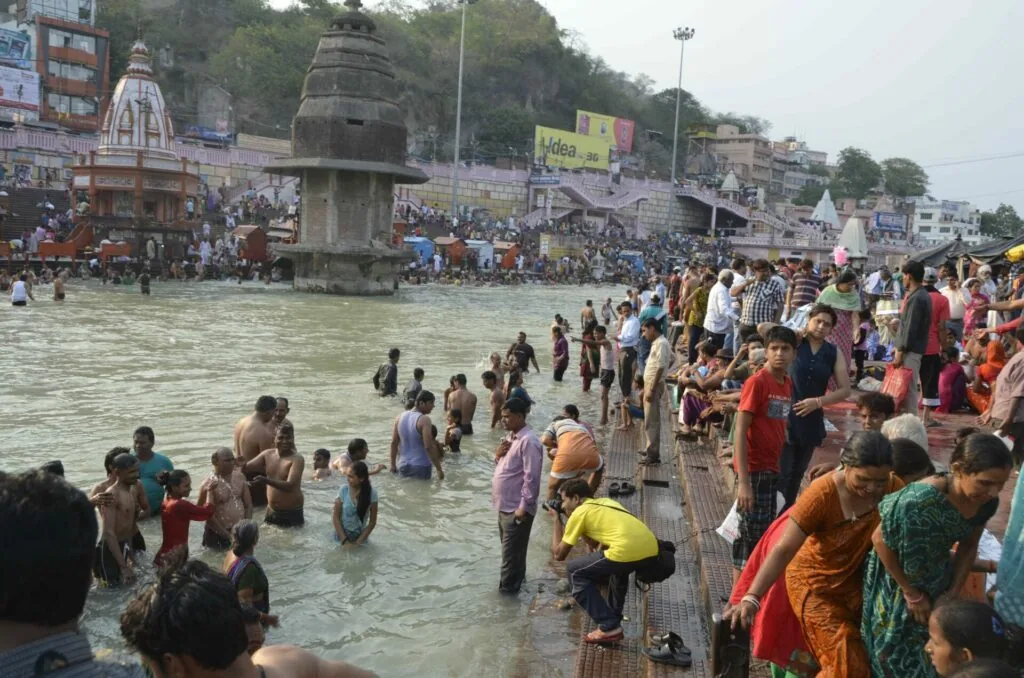 Large crowd of people bathing in a river with temples and buildings lining the banks