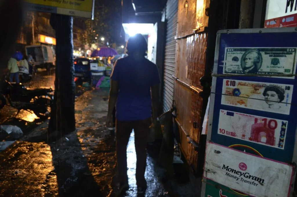 Person standing in narrow alley at night with currency exchange posters visible on brick wall