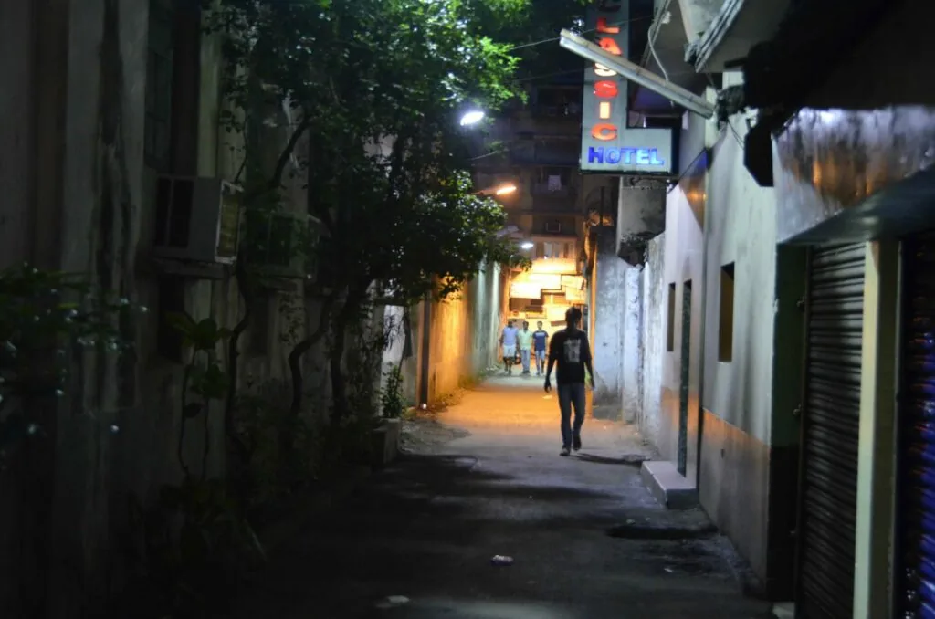 Nighttime urban alley with person walking, lit storefronts, hotel sign, and residential buildings