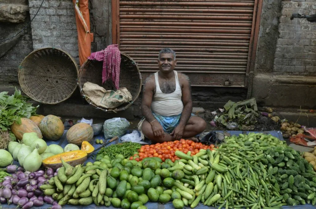 Vegetable vendor sitting behind display of fresh produce including tomatoes, beans, cucumbers, and leafy greens at street market
