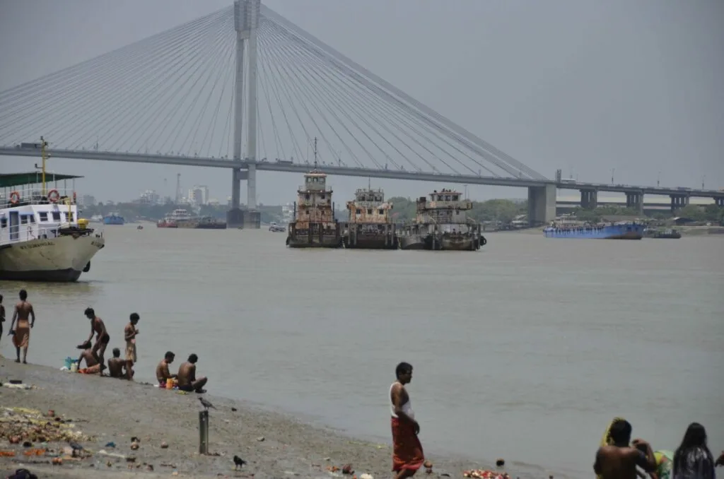 People wading and relaxing on a riverbank with tugboats and cargo vessels on the water beneath a cable-stayed bridge