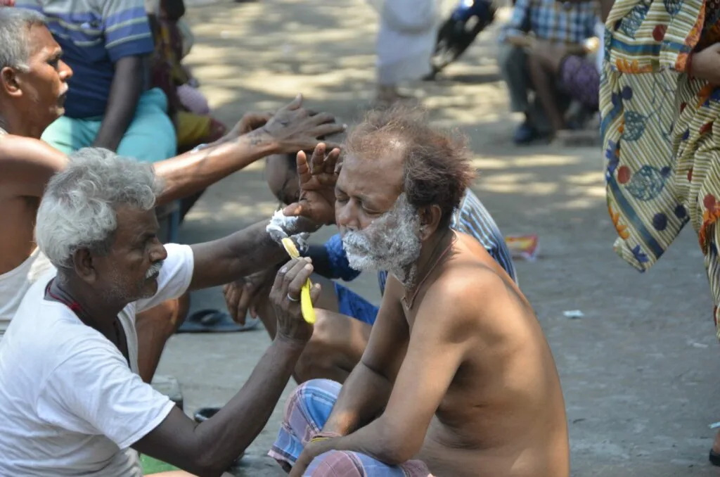 Elderly man with white beard receiving face paint or makeup applied by another man during a street gathering with onlookers in background
