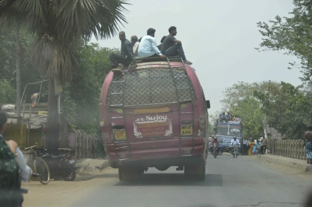 Four people sitting on top of an overcrowded maroon van with mesh cargo netting on a rural road lined with palm trees
