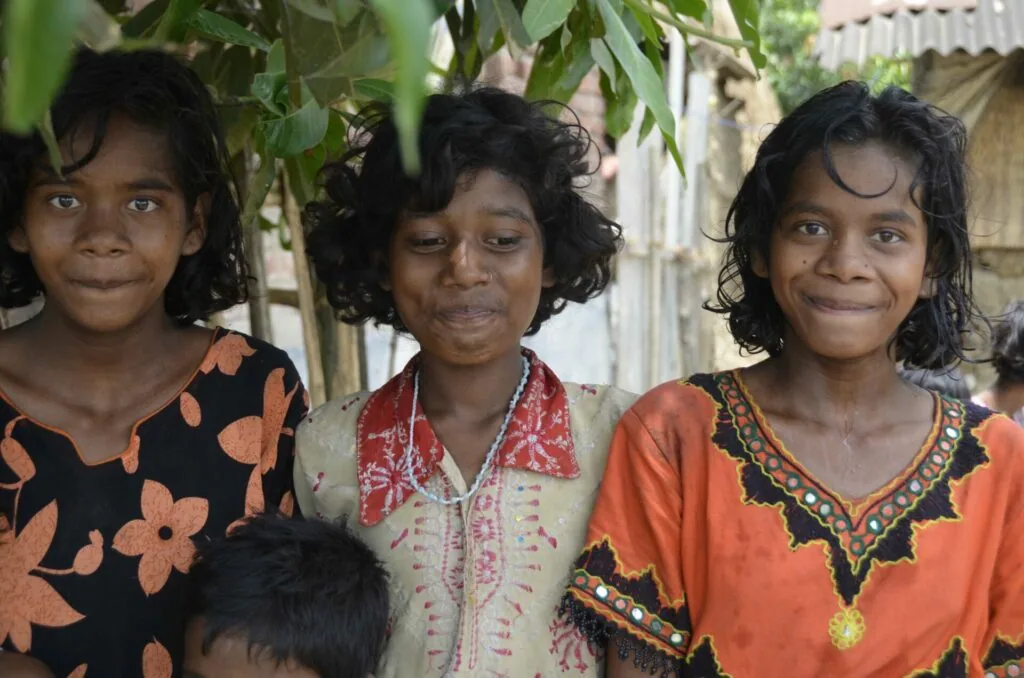 Three young girls smiling at camera under tree branches, wearing colorful patterned clothing