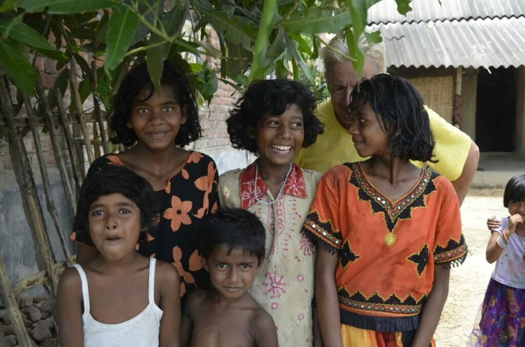 Group of children and an adult standing together outside a house with corrugated metal roof and banana plants nearby.