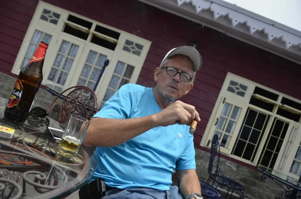 Man in blue shirt and cap sitting outdoors with beverage bottle and glass on table, red barn building with cream windows in background
