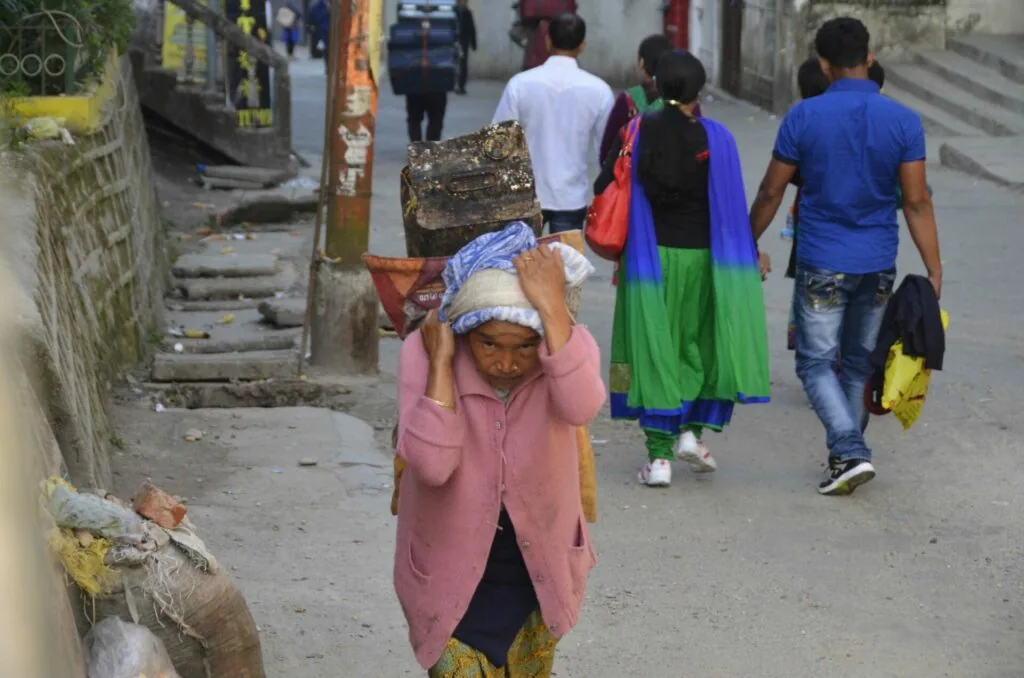 Woman carrying bundle of wood on head walking down narrow alley street with other pedestrians