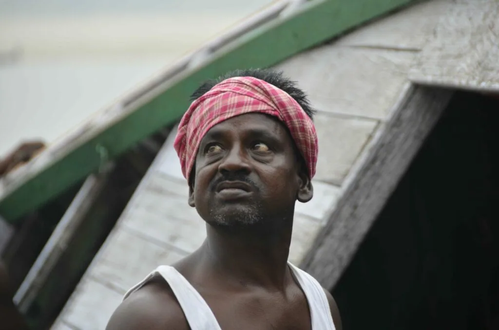 Man wearing pink checkered headwrap and white sleeveless shirt standing near weathered structure with green trim