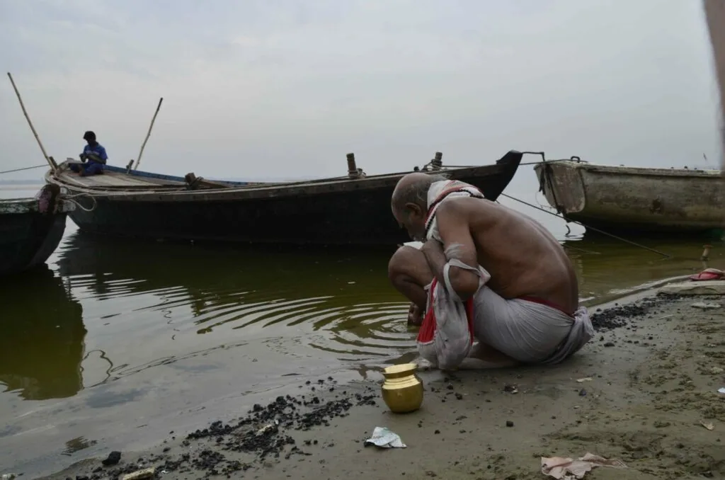 Man crouching on muddy riverbank near traditional boats moored in shallow water