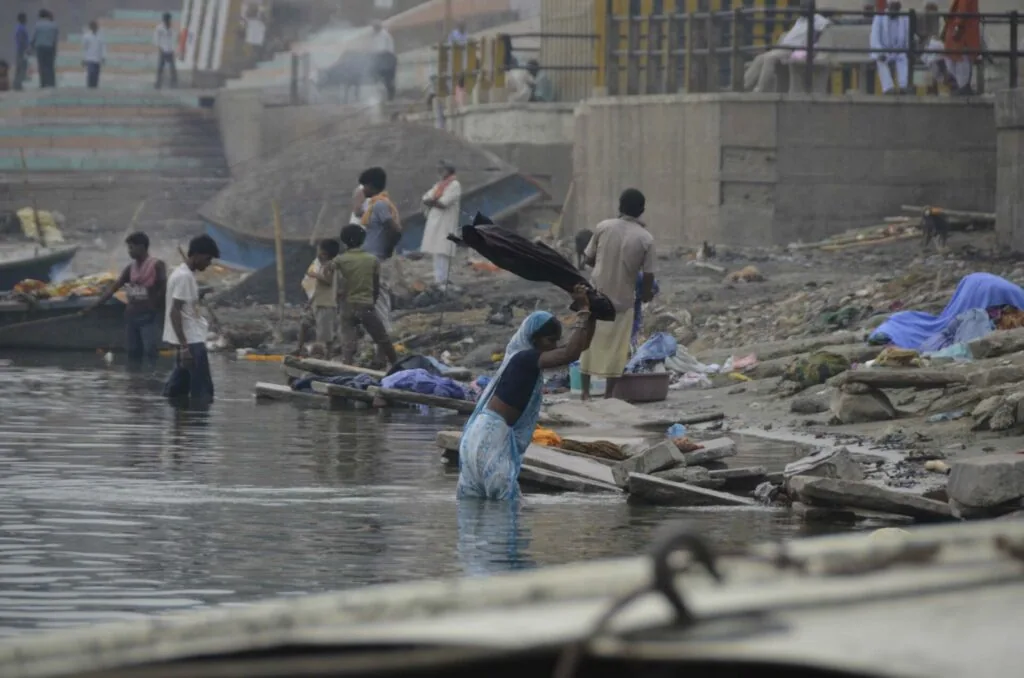 People washing clothes and bathing in a river with boats and buildings visible along a hazy waterfront