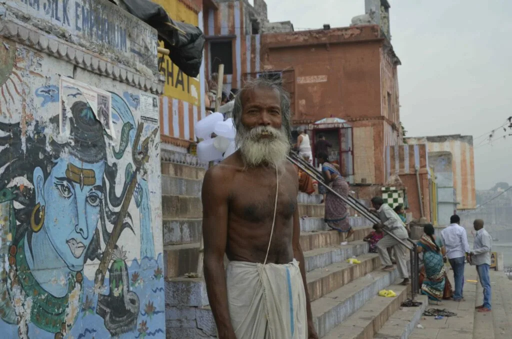 Elderly man with white beard holding staff stands before colorful mural artwork on waterfront buildings