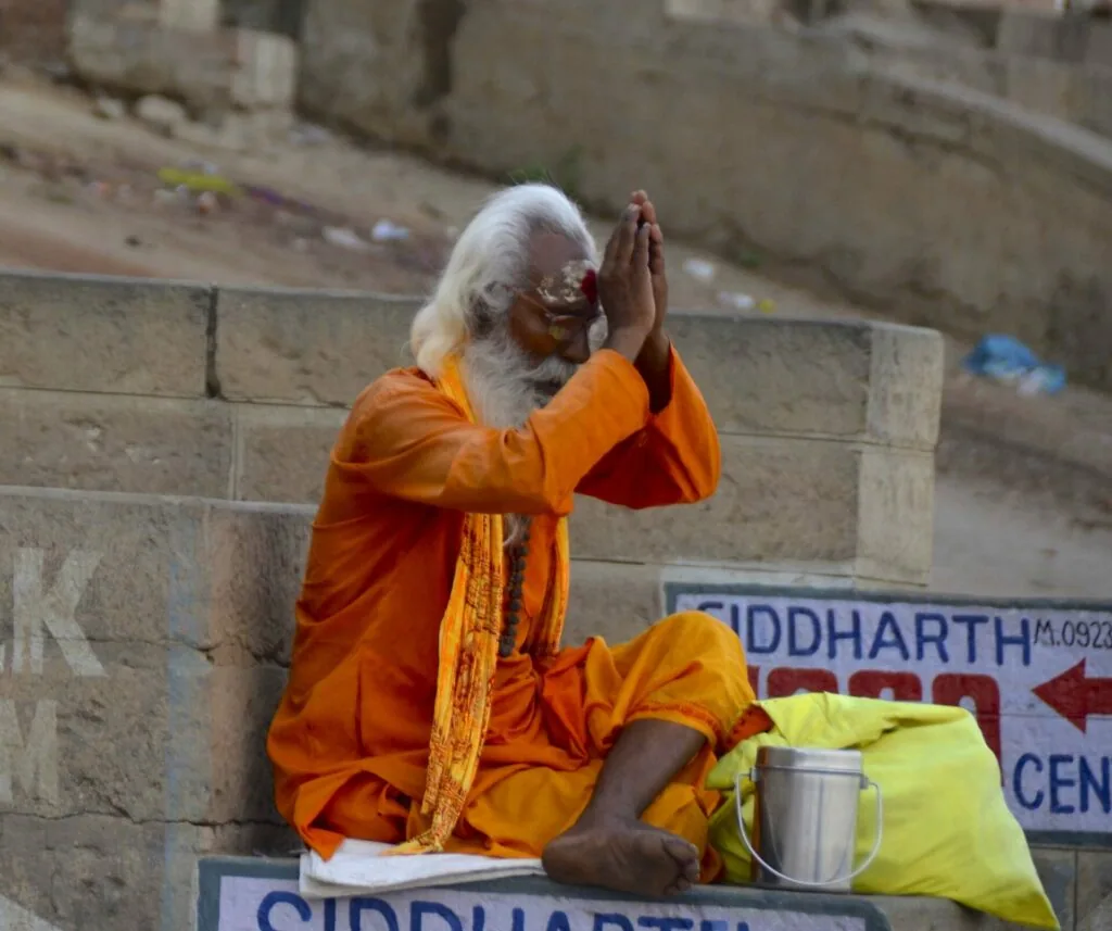 Elderly man with white hair and beard sitting on stone steps wearing orange robes with hands pressed together in prayer gesture