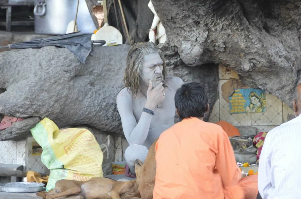 White statue of a figure with long hair in a meditative pose seated between large rocks, with a person in orange shirt viewed from behind in the foreground.