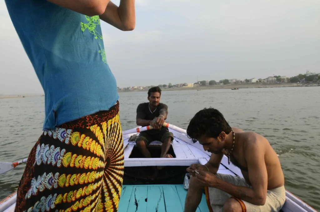 Three men in a small motorboat on a river with a colorful patterned fabric hanging on the left and residential buildings visible on the shoreline