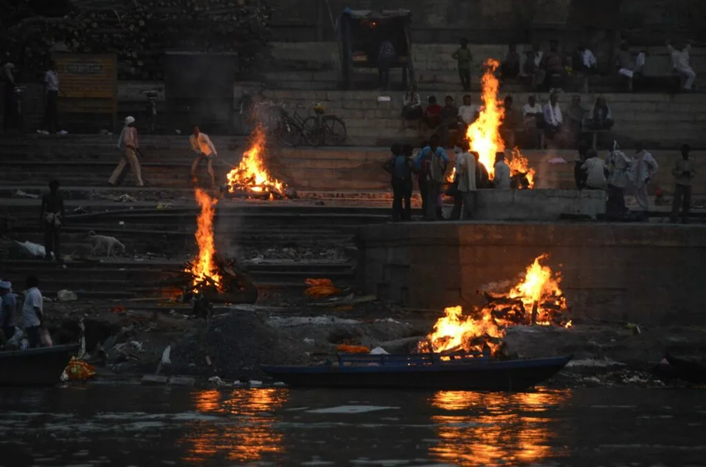 Multiple fires burning on stone steps and in a boat on a river at dusk with people gathered nearby
