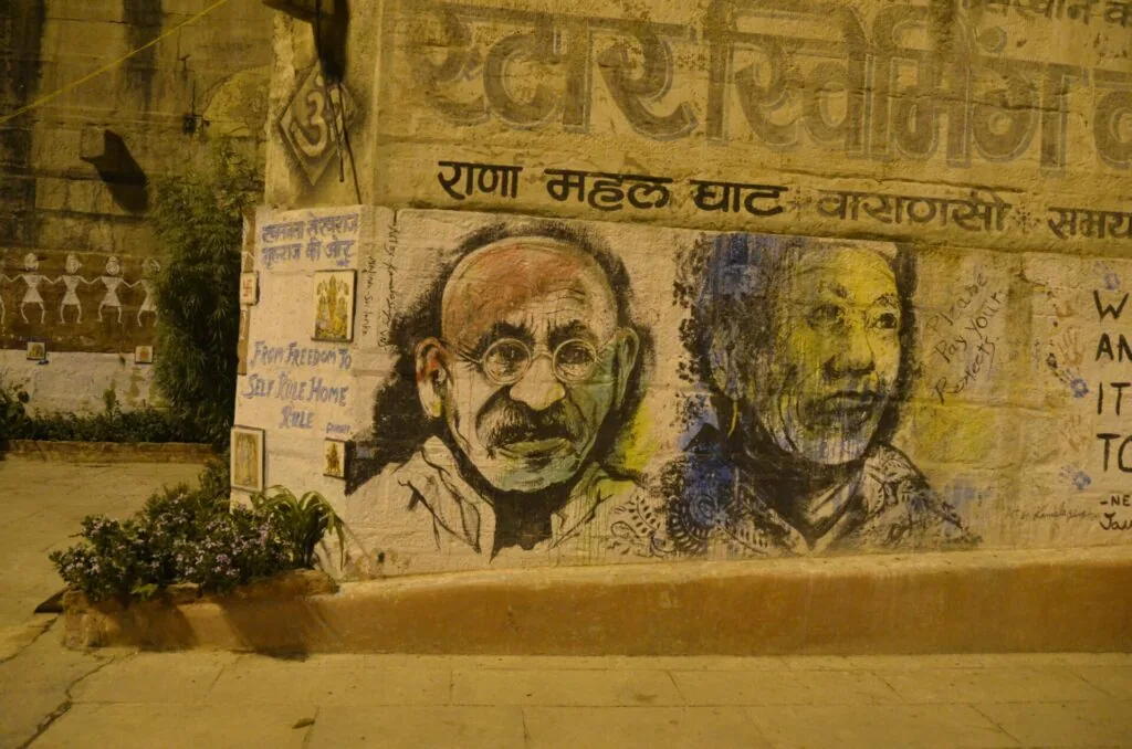 Street wall mural with two portrait drawings and Devanagari script text above, featuring weathered yellow and brown tones with planted vegetation at base