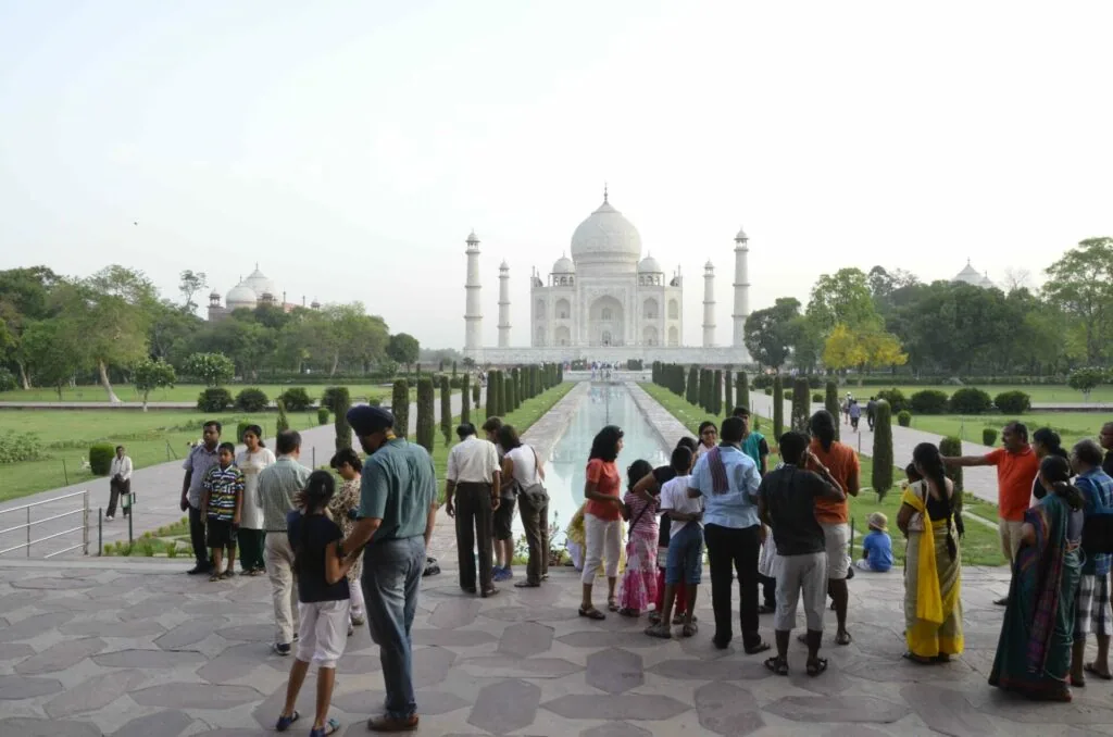 Tourists viewing a white domed mausoleum from a garden pathway lined with cypress trees, with manicured lawns on either side