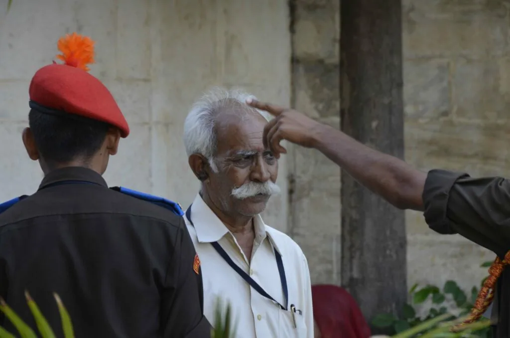 Elderly man with white mustache wearing white shirt with ID badge, interacting with uniformed person in red beret near stone wall