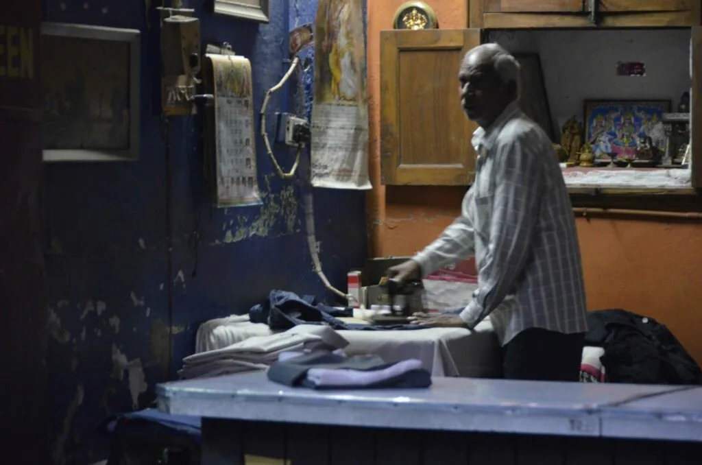 Elderly man in light striped shirt working at a table with textiles and materials in a workshop with blue and orange walls