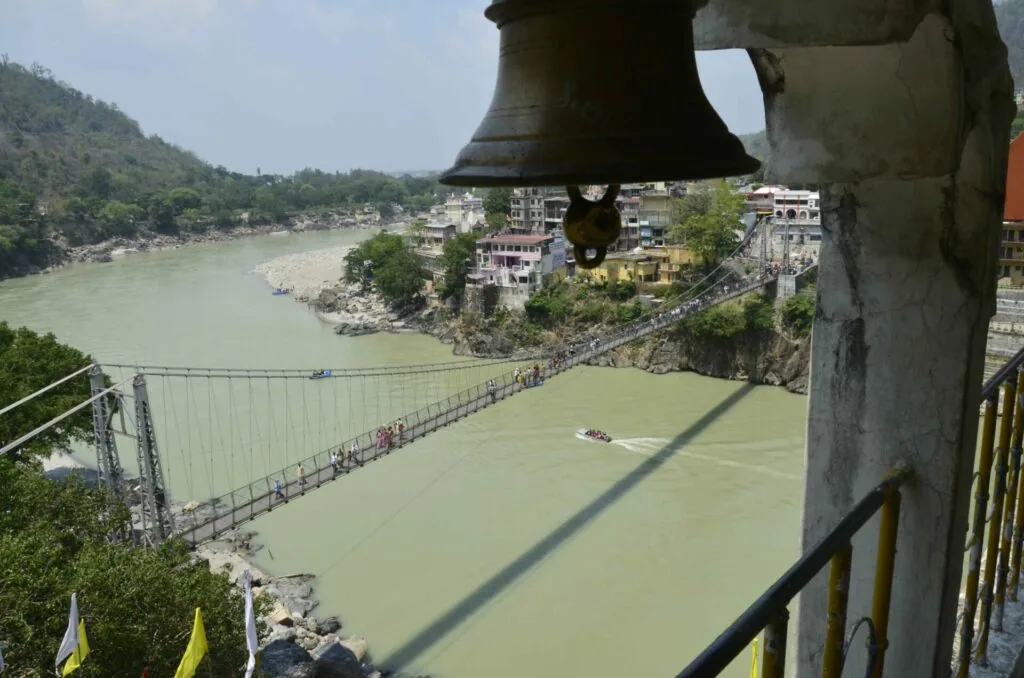 Large bronze bell hanging from stone structure overlooking suspension bridge crossing green river with hillside town beyond