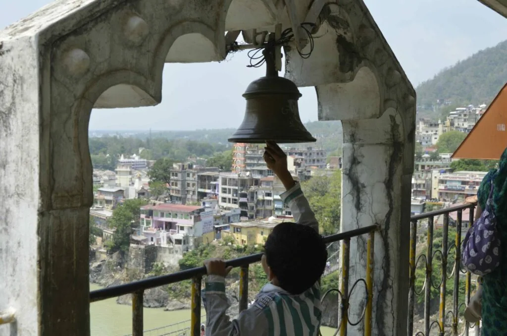 Person ringing ornate metal bell in stone archway overlooking valley with buildings and forested hills