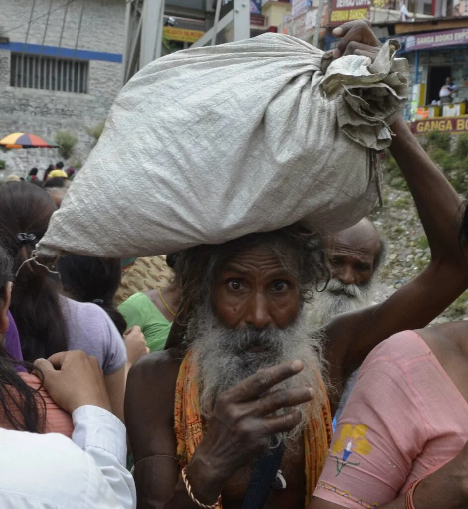 Elderly man with gray beard carrying large cloth sack on his head in crowded urban setting