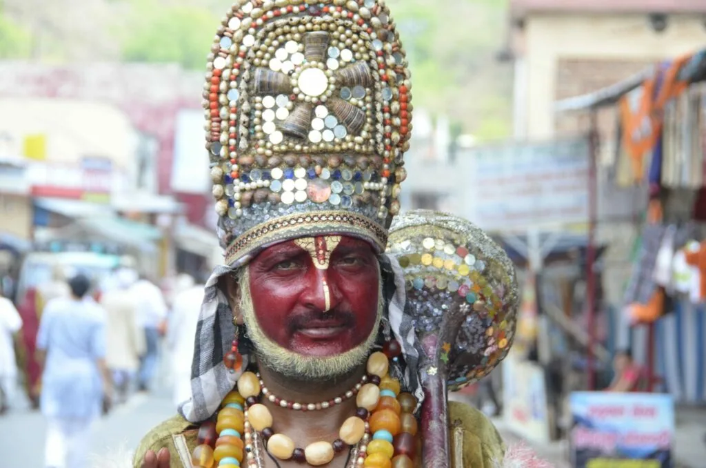 Person wearing traditional Indian ceremonial costume with tall beaded headdress, red face paint, and decorative beaded ornaments at a crowded street market