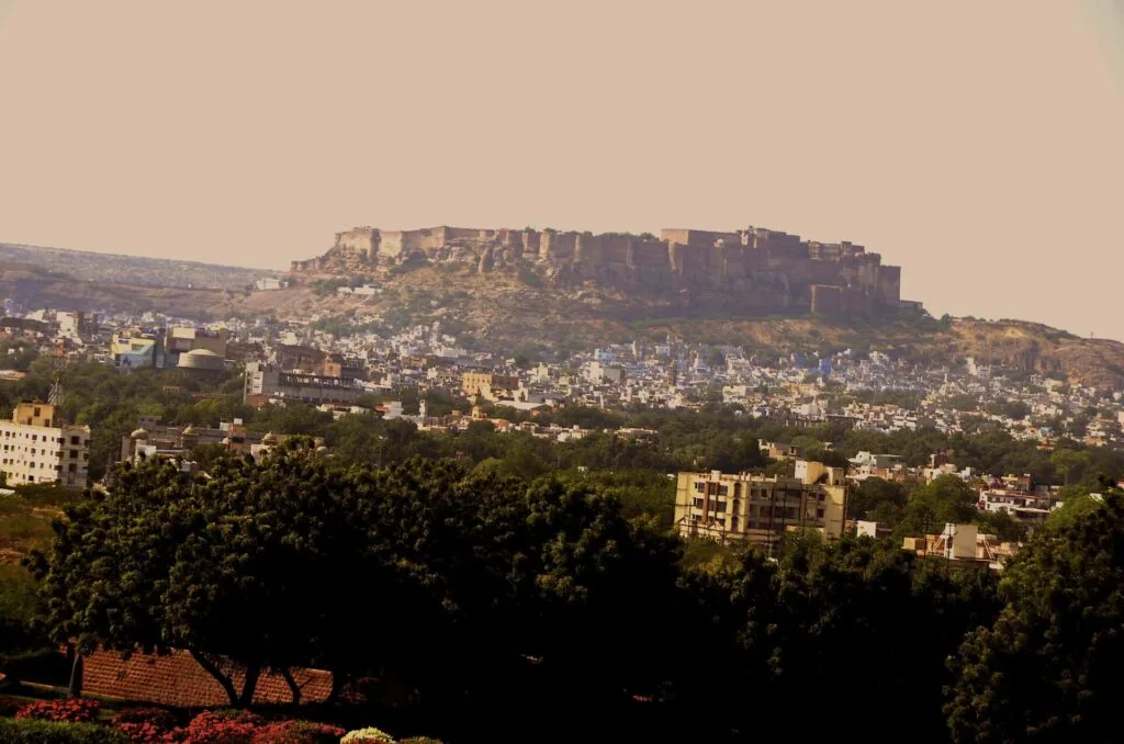 Fortified hilltop city with dense residential buildings sprawling across hillsides and green vegetation in foreground under hazy sky