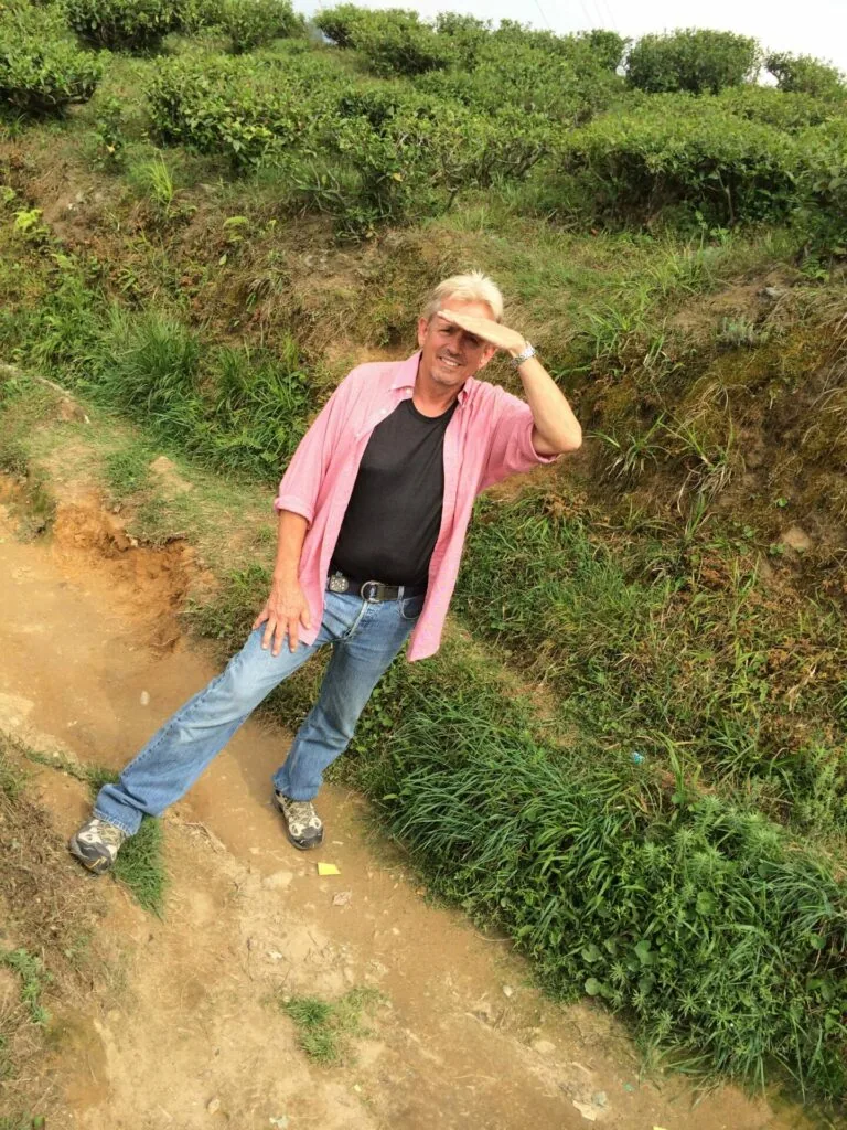 Man in pink shirt and straw hat standing on dirt path beside overgrown vegetation and hillside