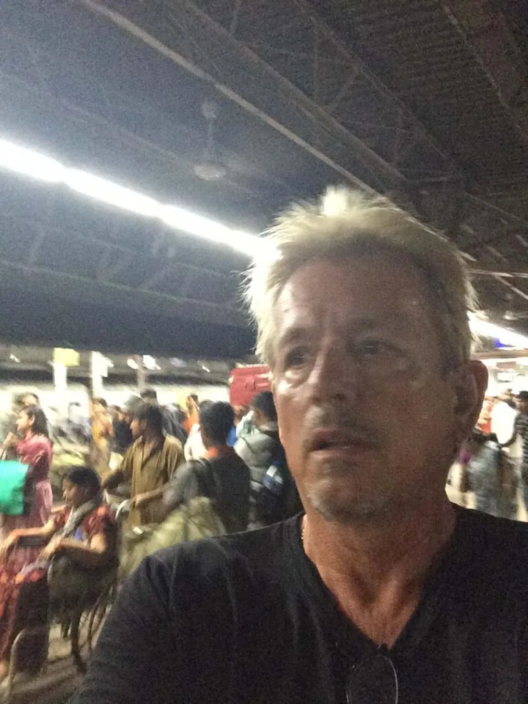 Man with light-colored hair in dark shirt at crowded indoor event under fluorescent lighting