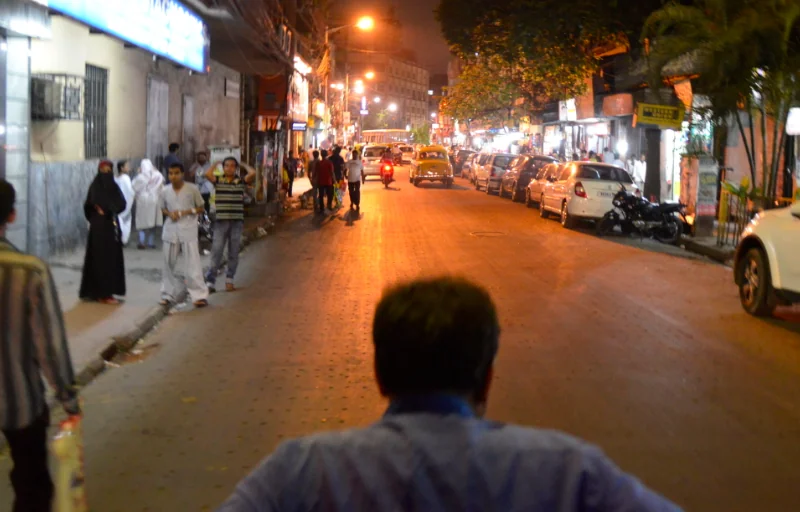 Person standing on illuminated nighttime street with pedestrians, parked vehicles, and storefronts