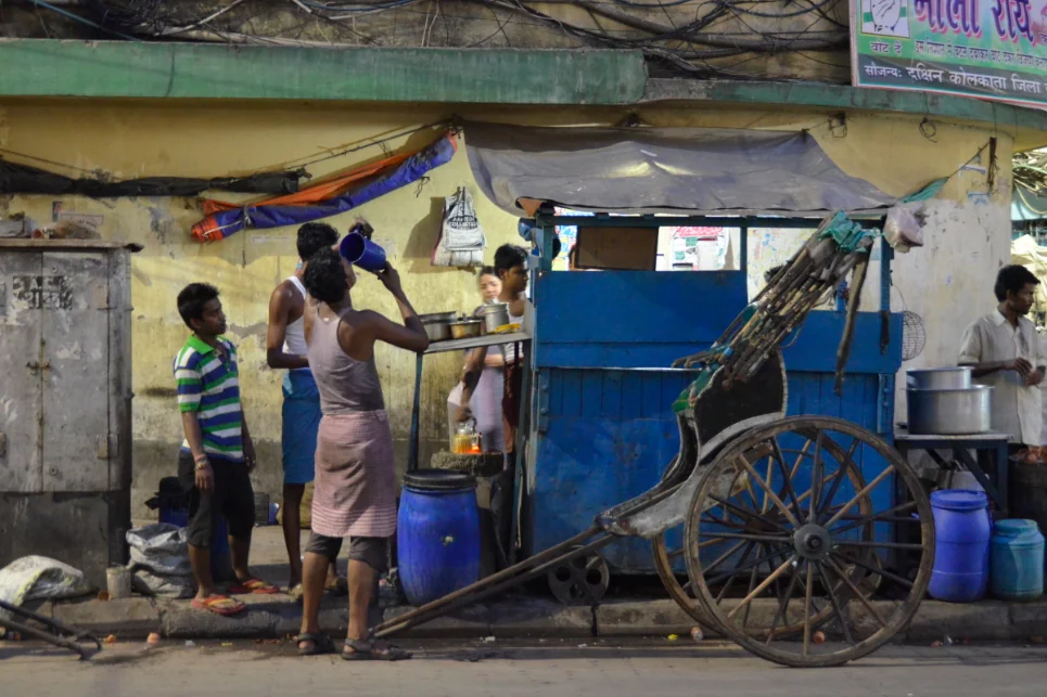 Street food vendor stall with yellow walls and blue counter, customers waiting, hand cart with large wheel in foreground