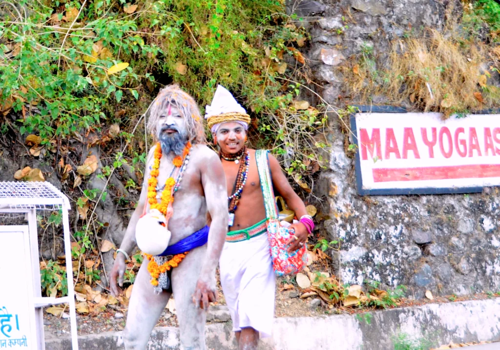 Two men in traditional Indian religious attire with body paint and garlands posing against a weathered stone wall with vegetation and a sign reading Maa Yoga.