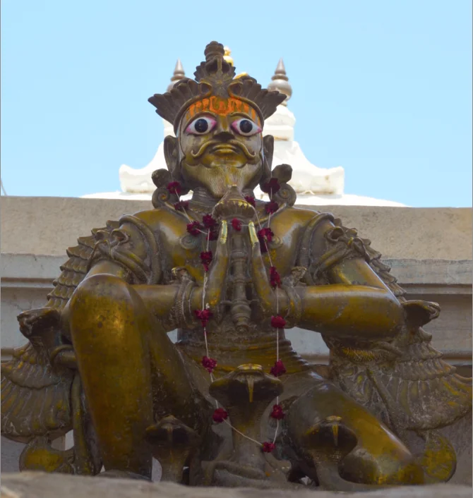 Bronze statue of a deity with ornate crown and decorative features, adorned with purple flower garlands, against clear sky.