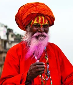 Man with long purple beard wearing red turban and red robes, holding prayer beads