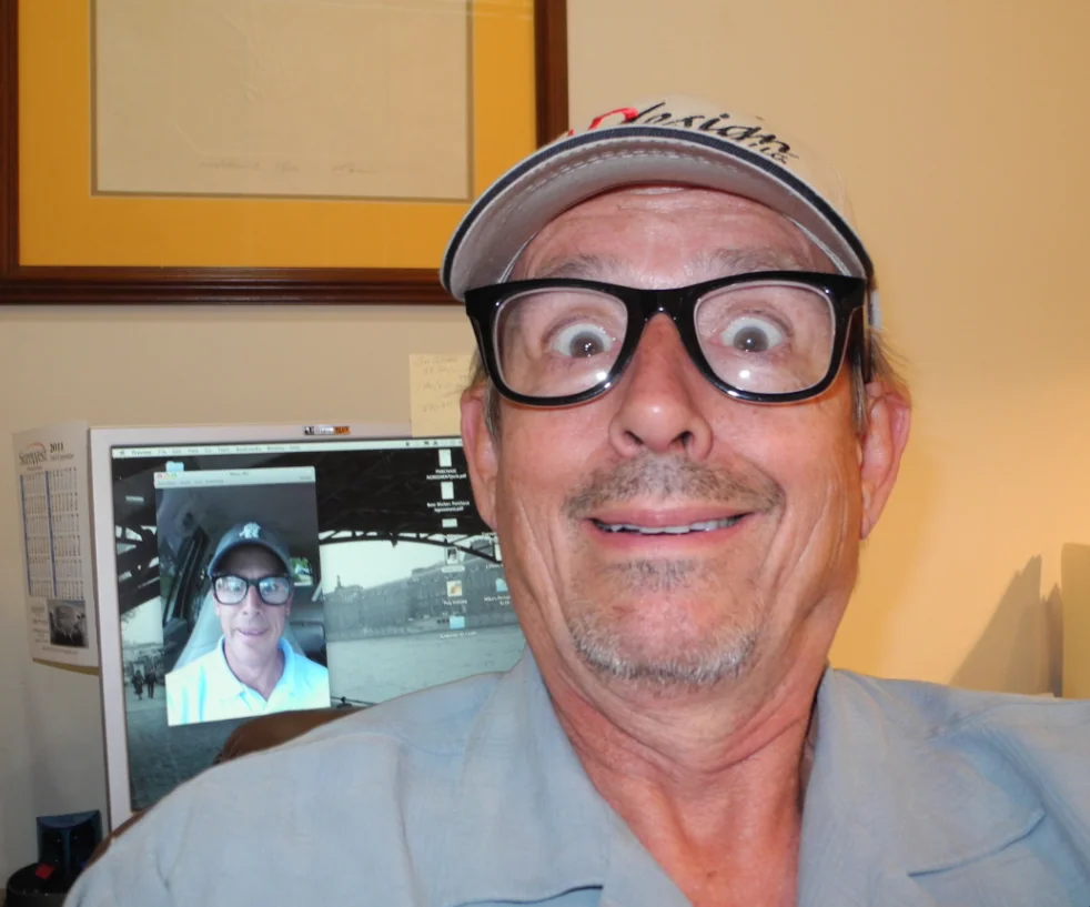 Man wearing glasses and cap smiling at camera with computer monitor showing video call in background