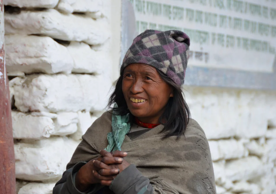 Woman wearing a purple plaid hat and gray shawl smiles while sitting against a white brick wall