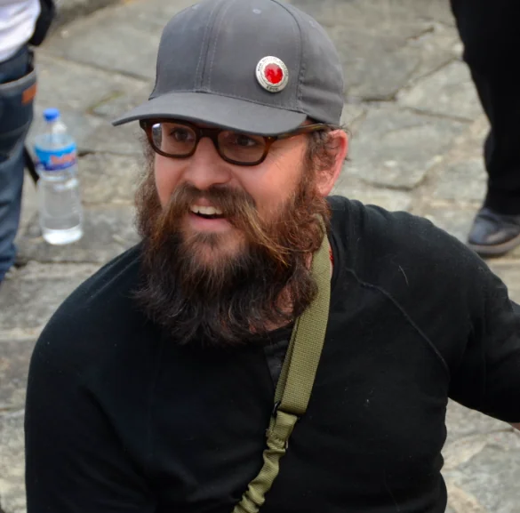 Man with long dark beard wearing glasses, gray cap, and black jacket at outdoor gathering
