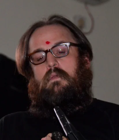 Man with long dark hair and beard wearing glasses and black jacket, holding microphone