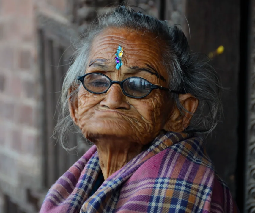 Elderly woman wearing glasses and a purple plaid shawl, with a blue bindi on her forehead