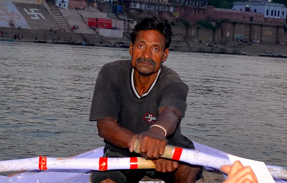 Man holding oar in small boat on river with historic waterfront buildings behind him