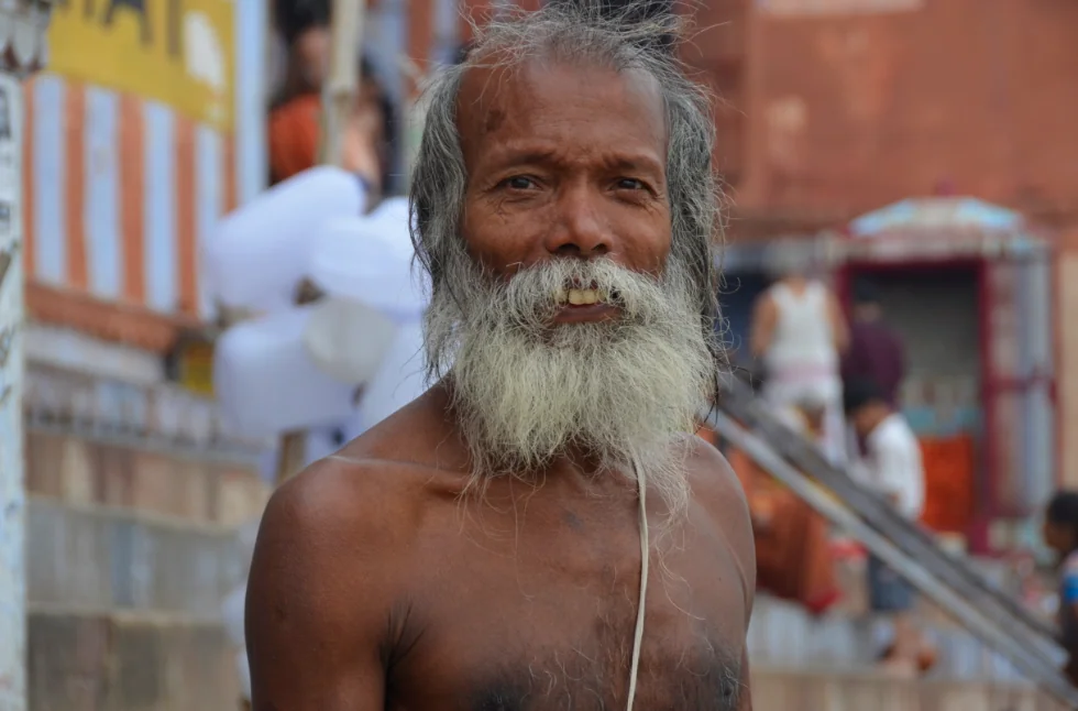 Elderly man with long white beard in outdoor temple setting with brick architecture and people in background