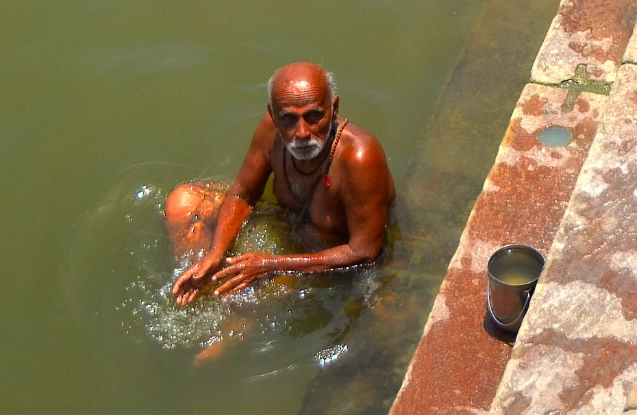 Older man with white beard bathing in greenish water beside brick ghat steps