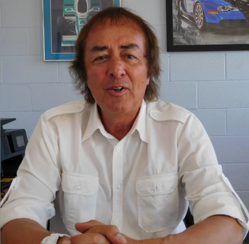Man with shoulder-length brown hair wearing white collared shirt sitting indoors with car artwork on wall behind him